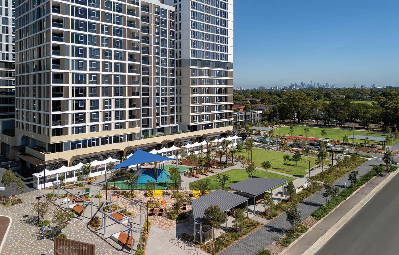 A modern high-rise building overlooks a landscaped outdoor area with a playground, shaded seating, walking paths, and a sports field under a clear blue sky. Description generated by AI