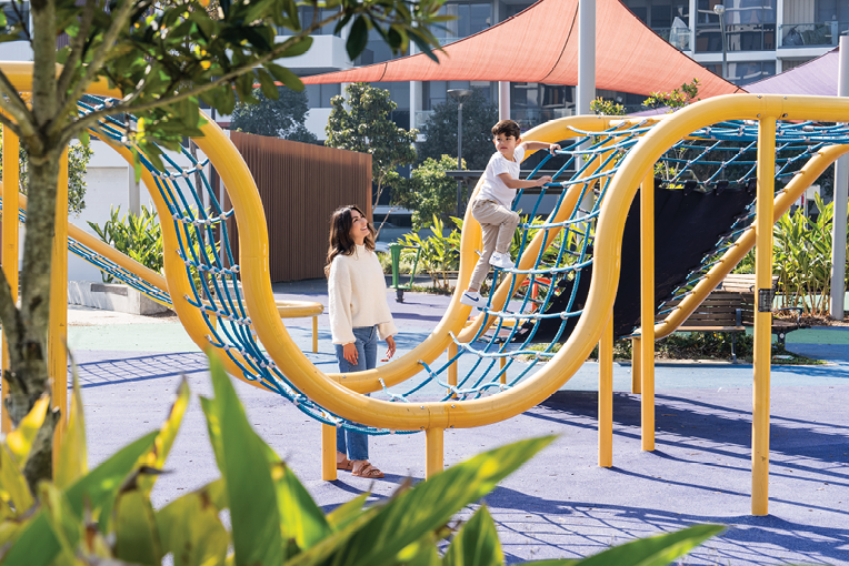 A person in a white sweater watches another person climbing a yellow playground structure with blue netting on a sunny day. Description generated by AI