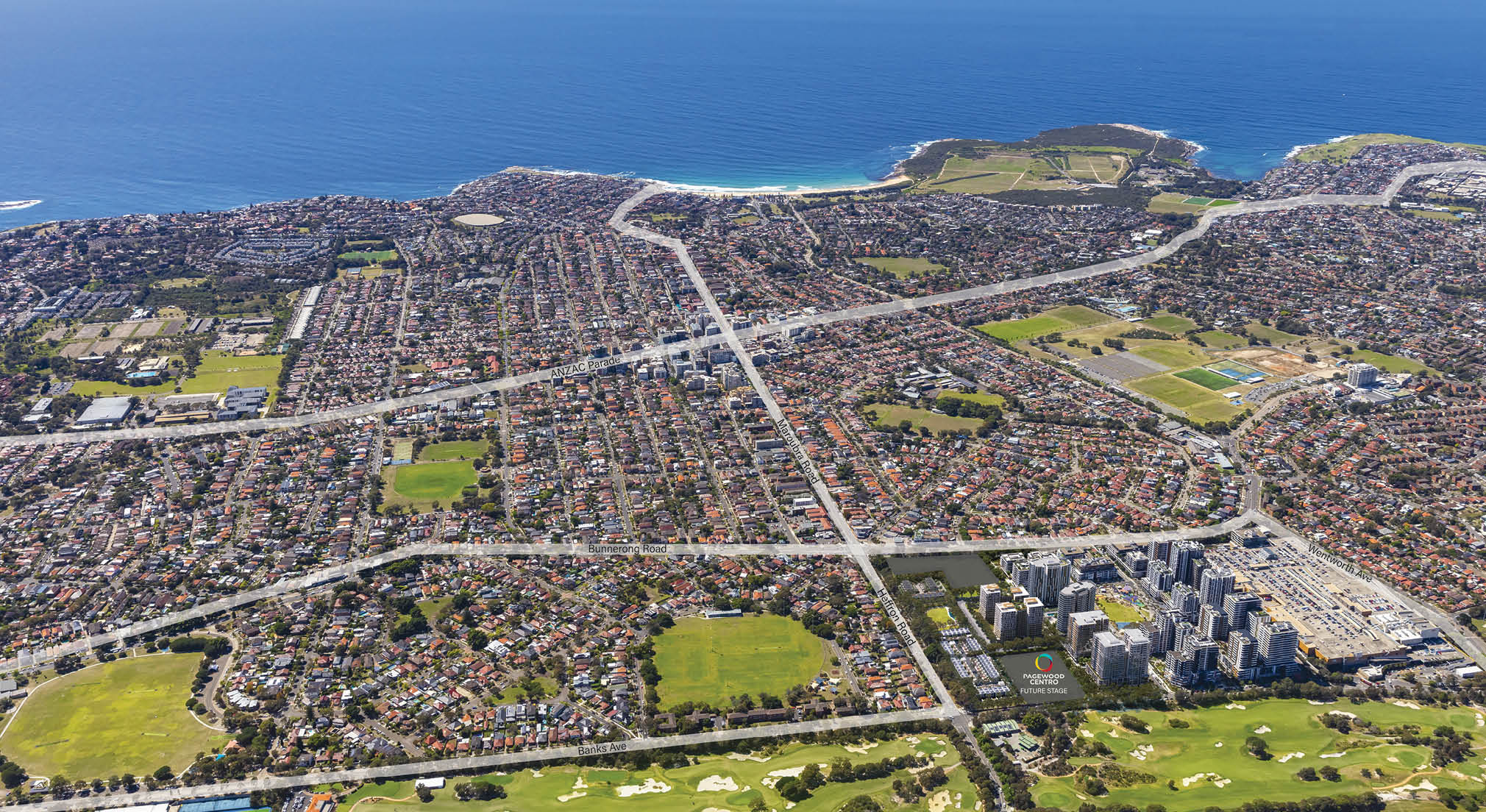 A coastal suburban area with densely packed houses, green parks, a cluster of tall buildings, and the ocean in the background. Description generated by AI