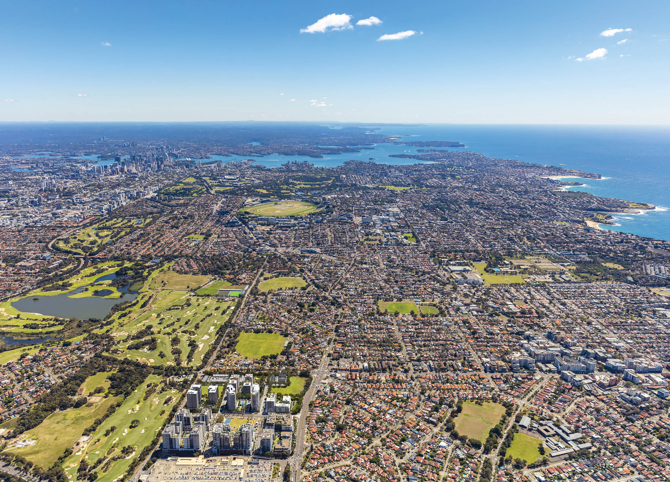 A sprawling urban landscape with dense residential and commercial buildings, patches of green parks and golf courses, and a coastline under a clear blue sky. Description generated by AI