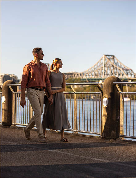Two adults holding hands walk along a waterfront path with a large bridge in the background under a clear sky. Description generated by AI