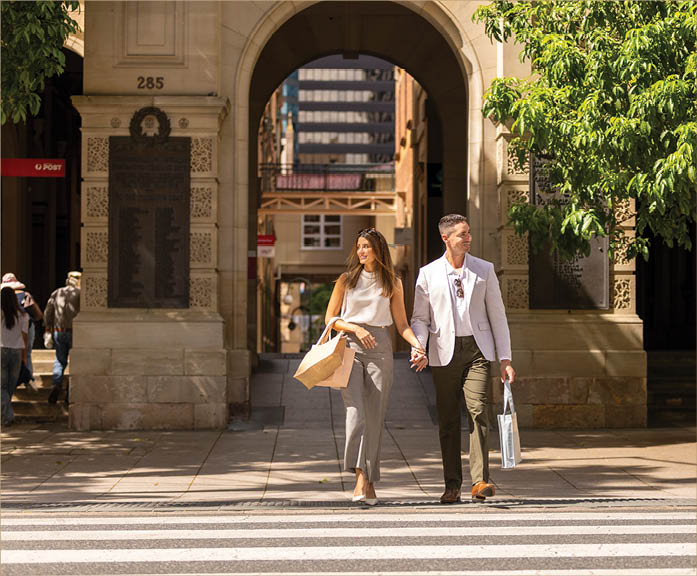 Two adults dressed in stylish clothing are walking hand in hand across a crosswalk in an urban area with trees and buildings in the background. Description generated by AI