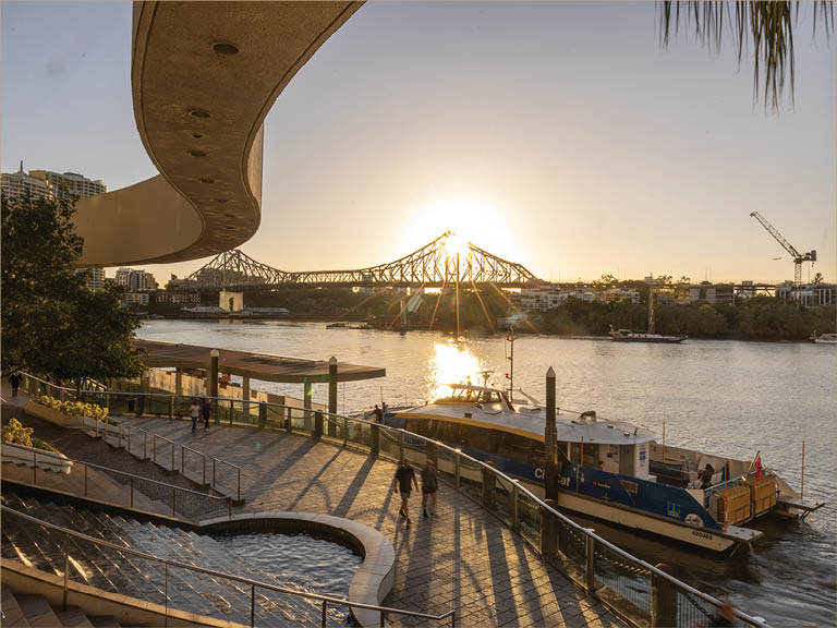 A river scene at sunset with a bridge in the background, a boat docked by a curved walkway, and people walking along the riverside path. Description generated by AI