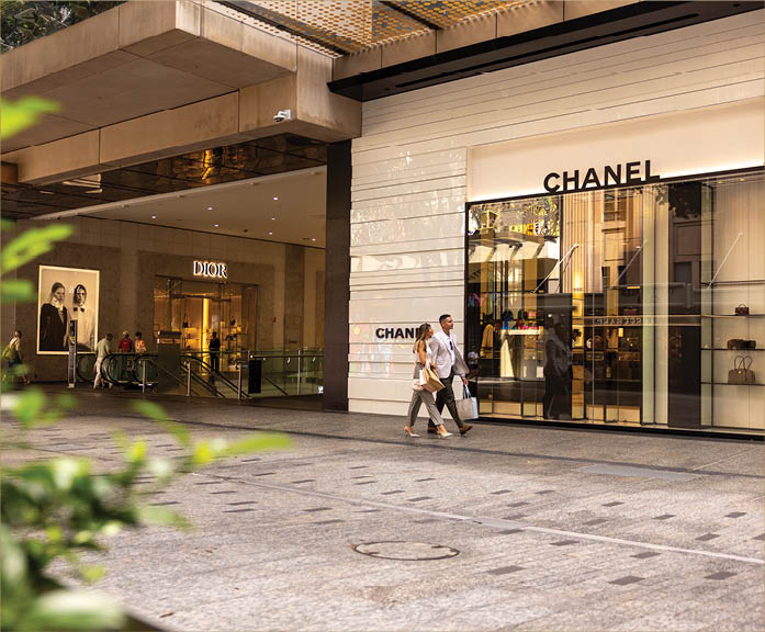 Two adults walk past luxury storefronts of Chanel and Dior in a modern outdoor shopping area. Description generated by AI