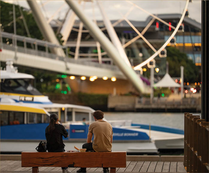 Two people sit on a wooden bench by the water with boats and a large Ferris wheel in the background during dusk. Description generated by AI