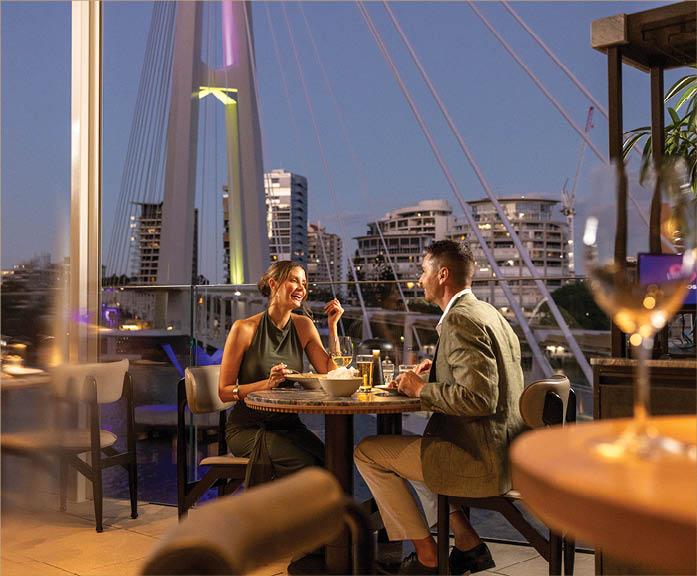 Two adults dressed in semi-formal attire are sitting at a table enjoying a meal and drinks in a modern restaurant with large windows overlooking a cityscape and illuminated bridge at dusk. Description generated by AI