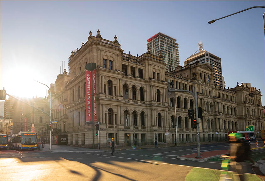 A historic stone building with arched windows and a red Griffith University banner stands at a sunlit city intersection with blurred people and vehicles in motion. Description generated by AI