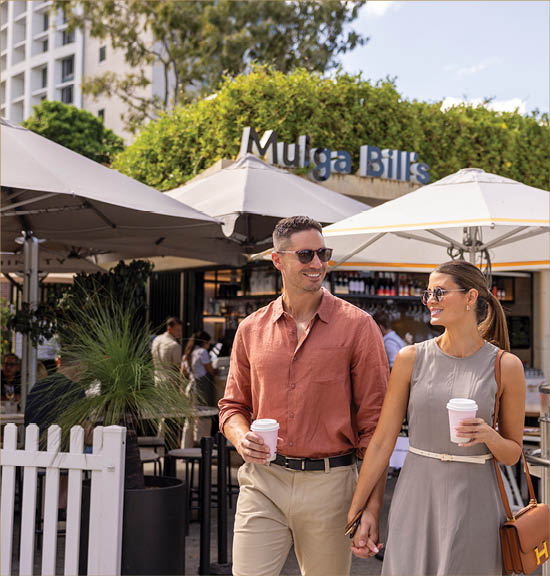 Two adults wearing sunglasses and casual clothing hold hands and carry coffee cups while walking outside a caf with umbrellas and greenery in the background.  Description generated by AI