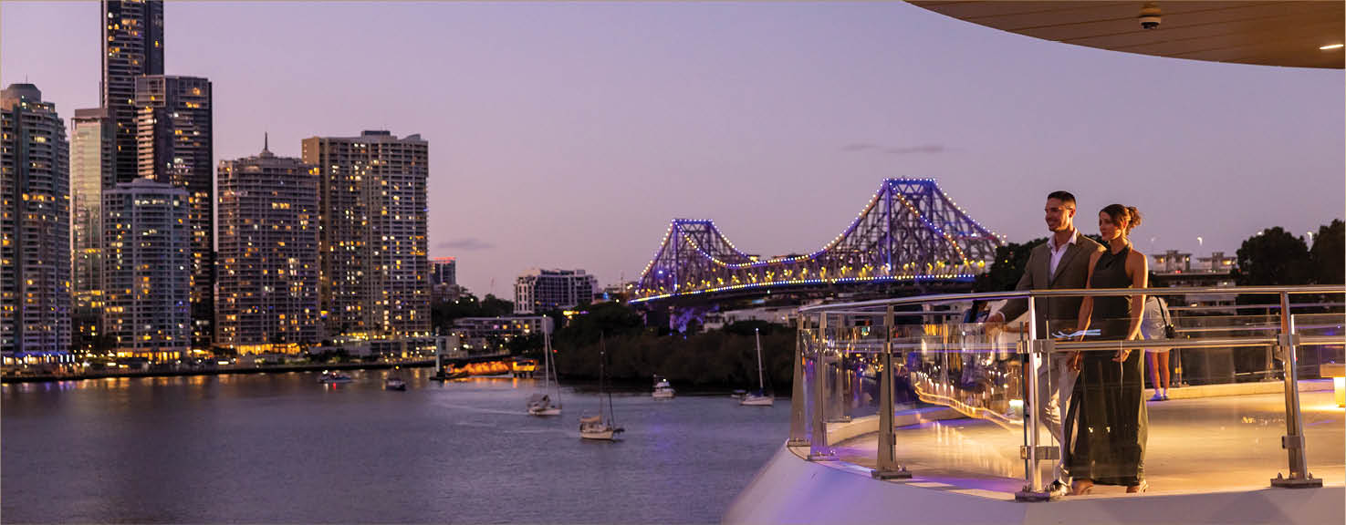 Two people dressed in formal attire stand on a balcony overlooking a river with a city skyline and illuminated bridge at dusk. Description generated by AI