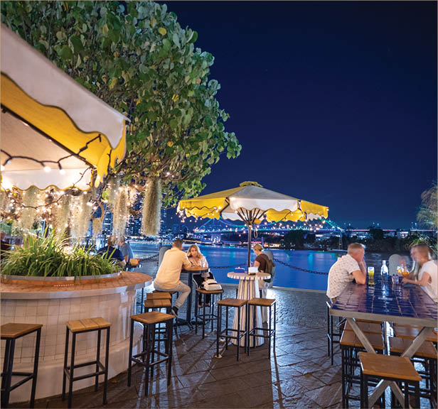 People sit at outdoor tables under string lights and umbrellas at night with a cityscape in the background. Description generated by AI
