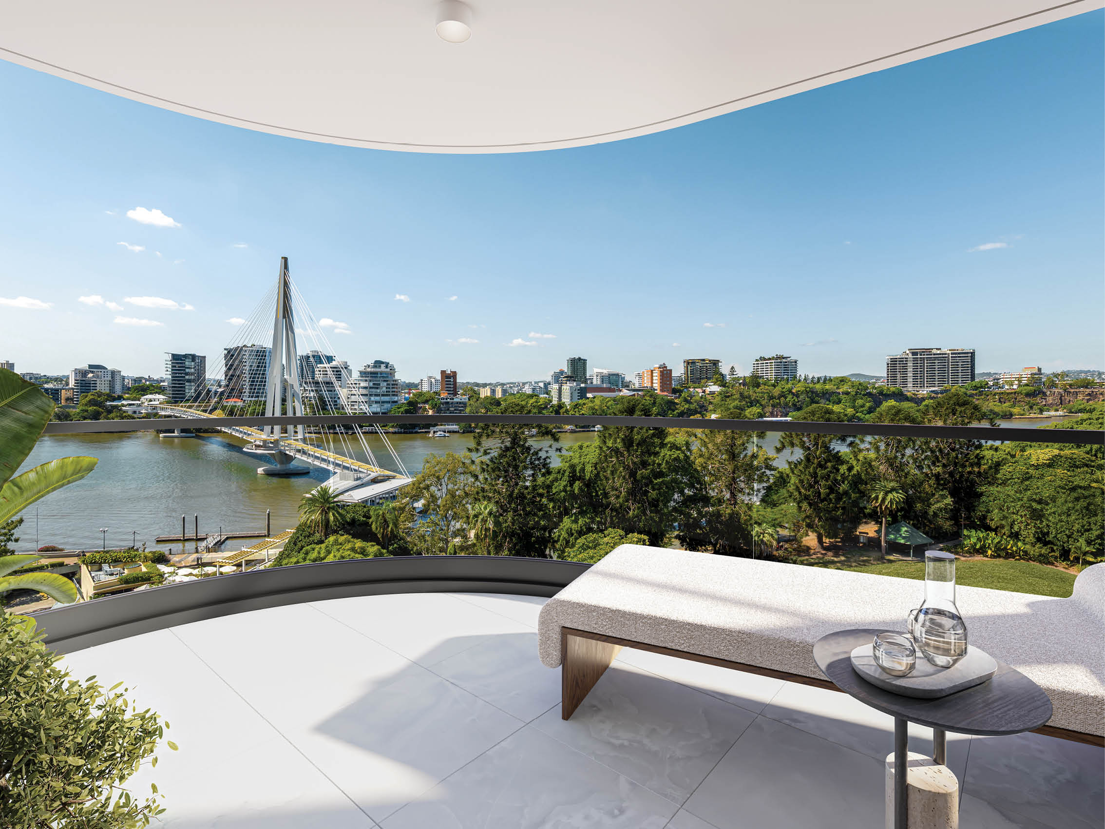 A modern balcony with a lounge chair and small table overlooks a river with a distinctive cable-stayed pedestrian bridge and a city skyline in the background under a clear blue sky. Description generated by AI