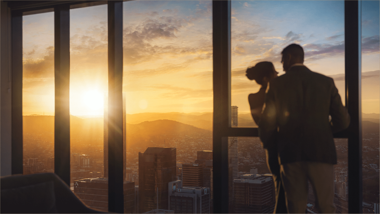 Two adults stand close together by large windows overlooking a cityscape at sunset with warm light filling the room. Description generated by AI