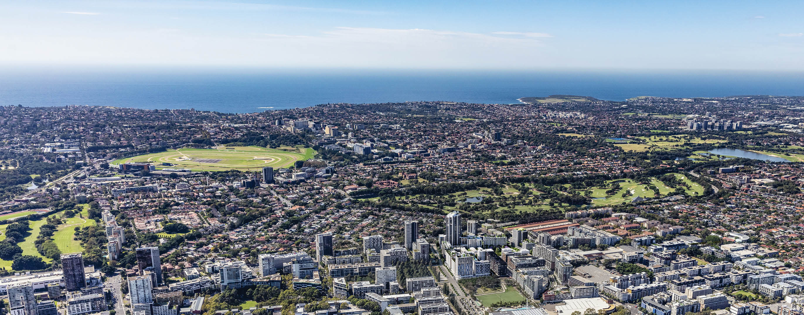 A sprawling urban landscape with numerous buildings, green parks, and a distant coastline under a clear sky. Description generated by AI