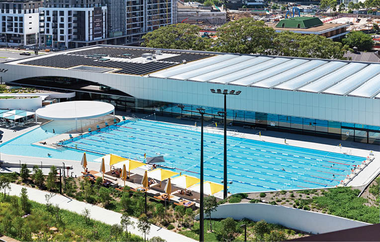 An outdoor swimming pool with lanes is surrounded by a modern building, lounge chairs with umbrellas, and greenery in an urban setting. Description generated by AI