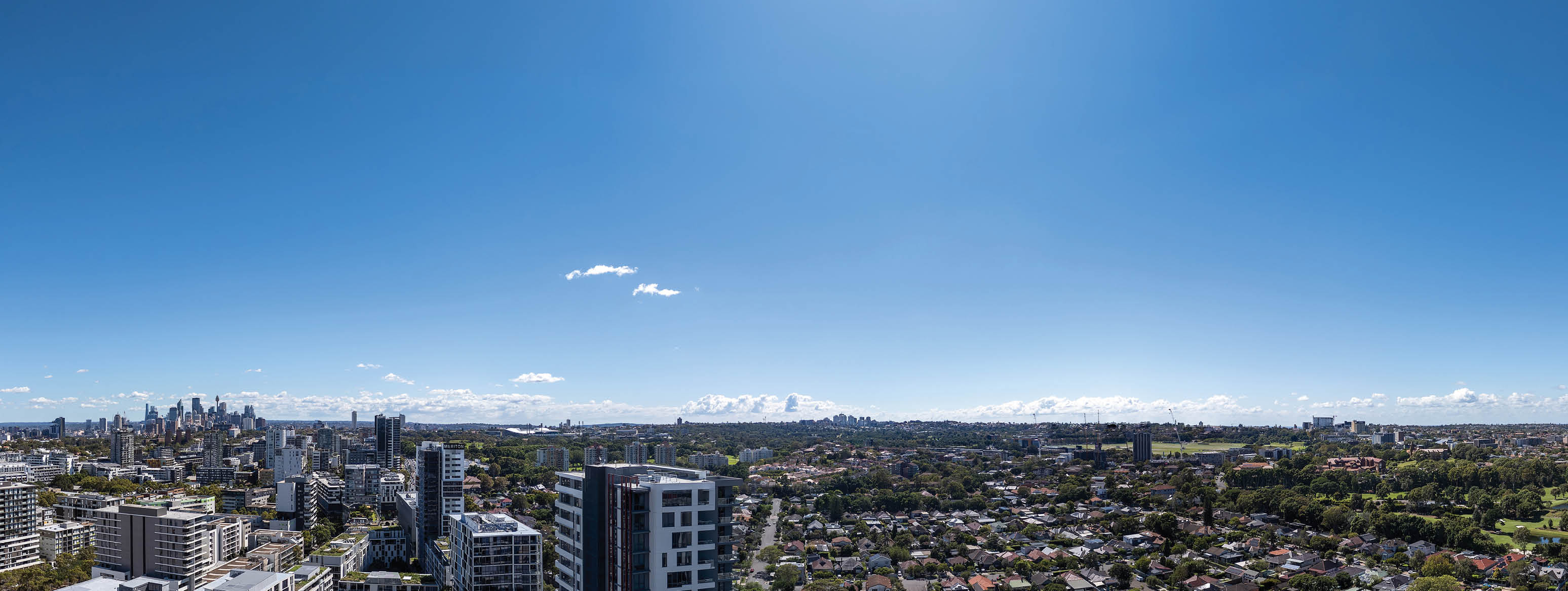 A clear blue sky stretches over a cityscape with a mix of tall buildings and residential houses surrounded by greenery. Description generated by AI